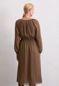 Bruine jurk gemaakt van een semi-doorzichtig materiaal, met een elastische taille, lange mouwen en een geometrisch wit patroon overal.