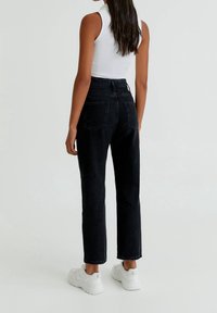 Femme portant un jean noir taille haute, un top blanc sans manches et des baskets blanches épaisses, debout devant un fond uni clair.