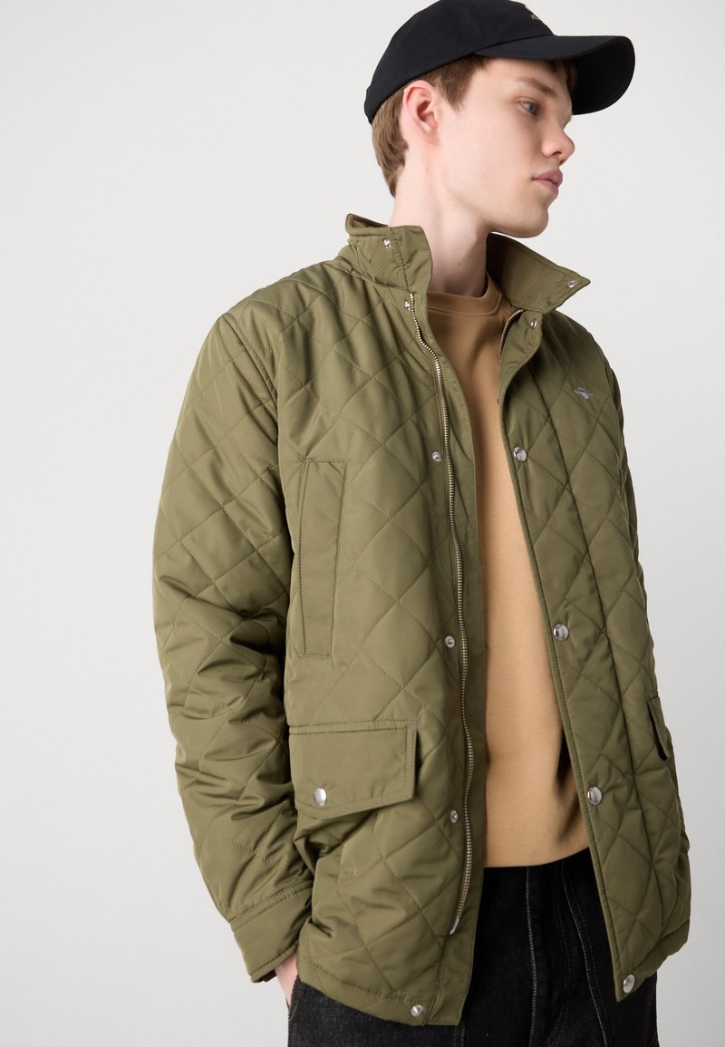 Zielonooliwkowa pikowana kurtka z wysokim kołnierzem, napami, bocznymi kieszeniami i gładką teksturą, noszona na beżowej bluzie.