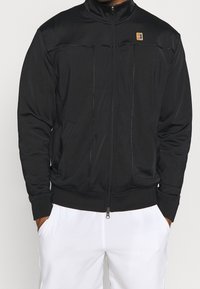 Svart zip-up jacka med ståkrage, två frampockets och subtil sömdetaljering. Tillverkad av ett smidigt, lätt material.