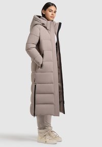 Langer Puffer-Mantel in Hellbeige mit Kapuze, ausgestattet mit einer gesteppten Oberfläche, zwei seitlichen Reißverschlusstaschen und einem kontrastierenden schwarzen Reißverschluss.