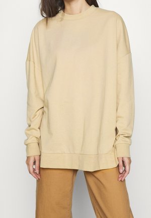 Personne portant un sweat-shirt beige ample à manches longues et un pantalon marron clair, debout devant un fond uni.