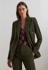 Groen geruite blazer met een dubbele knoopsluiting, gouden knopen en bijpassende broek. Daaronder een bordeauxkleurige bloemen-top met franje.