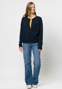 Cardigan albastru închis cu guler rotund, închidere cu butoane în față și detalii brodate, asortat cu blugi evazați de culoare albastru deschis și adidași albi.