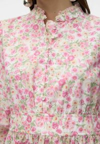 Närbild av en blommig klänning med rosa och gröna blommor, runda tygklädda knappar och en volangkrage, bärs tillsammans med stora runda silverörhängen.