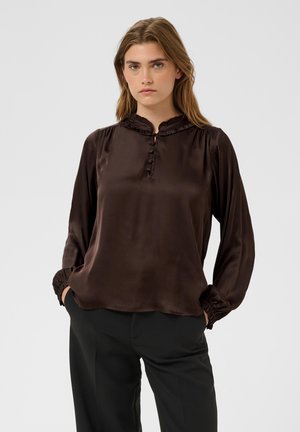 Femme aux cheveux châtain clair portant un chemisier marron foncé à manches longues avec des boutons et un pantalon noir, debout les mains dans les poches.