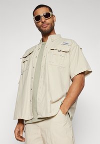 Camicia beige a maniche corte con tasche sul petto e chiusura a bottoni, indossata sopra una maglietta verde chiaro. Accompagnata da occhiali da sole.
