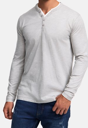 Homme portant un t-shirt henley à manches longues gris clair avec trois boutons et un jean bleu foncé déchiré, main posée près de la poche.