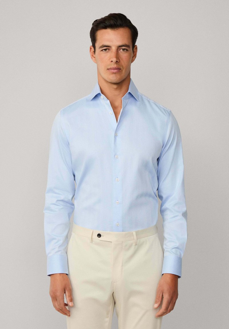 Muškarac u svijetlopljoj košulji s dugim rukavima i bež hlačama, stoji s opuštenim rukama naslonjenim na običnu sivu pozadinu.