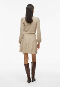 Maglione beige lavorato a maglia con maniche a sbuffo, abbinato a una gonna a campana coordinata. La modella indossa degli stivali alti in pelle marrone.