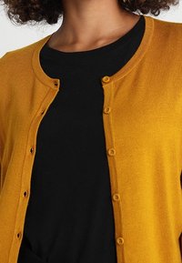Kvinna som bär en senapsgul knäppt kofta över en svart rundhalsad tröja, med lockigt hår delvis synligt.