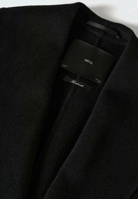 Manteau noir en mélange de laine avec un col à col en V profond. Présente une étiquette indiquant "MNG" et "Fait main", mettant en valeur une finition lisse et texturée.