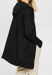 Svart hoodie-kardigan med långa ärmar, tillverkad av lättviktsmaterial. Har framfickor och en avslappnad passform, med ribbad nederkant.