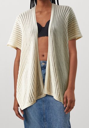 Kvinde iført åben cremefarvet strikket cardigan over sort bh og blå denimnederdel mod ensfarvet baggrund.