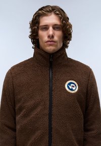 Άνδρας με σγουρά μαλλιά φοράει καφέ fleece μπουφάν μέχρι το λαιμό με επώνυμο πατς στο αριστερό στήθος, μπροστά σε απλό ανοιχτό φόντο.
