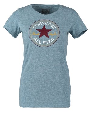 Osoba w jasnoniebieskim t-shircie Converse All Star, czarnych obcisłych spodniach i granatowych sneakersach, stoi z jedną ręką w kieszeni.