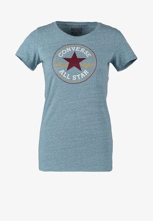 Osoba w jasnoniebieskim t-shircie Converse All Star, czarnych obcisłych spodniach i granatowych sneakersach, stoi z jedną ręką w kieszeni.