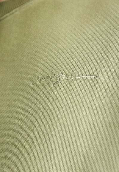 Tessuto a maglia verde oliva con fine costina e una texture sottile. Presenta un piccolo logo ricamato di colore chiaro.