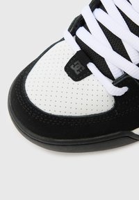 Ténis preto e branco com biqueira em couro branco perfurado, detalhes em camurça preta, sola espessa de borracha preta e atacadores planos brancos.