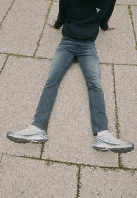 Person sitting on textured pavement with legs extended, wearing blue jeans, white and blue athletic shoes, and a black Barbour sweatshirt.