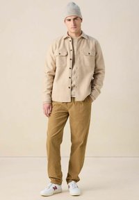 Veste chemise beige à boutons avec deux poches, associée à un pantalon en velours côtelé tan texturé et des basket blanches, surmontée d'un bonnet en tricot gris clair.