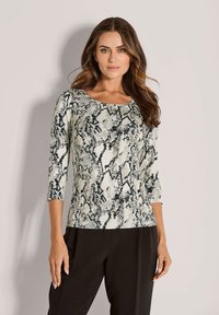 Frau mit welligem braunem Haar, die ein schwarz-weißes Oberteil mit Schlangenprint und drei Viertelärmeln sowie schwarze Hosen trägt, steht an einer hellen Wand.