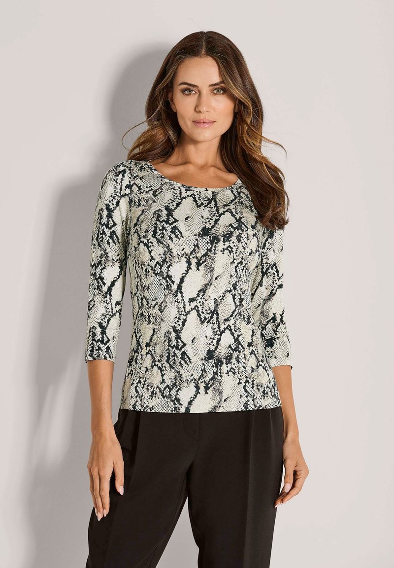 Frau mit welligem braunem Haar, die ein schwarz-weißes Oberteil mit Schlangenprint und drei Viertelärmeln sowie schwarze Hosen trägt, steht an einer hellen Wand.