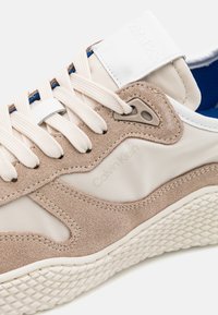 Baskets en suède beige et synthétique beige avec des accents en cuir blanc, doublure intérieure bleue et semelle en caoutchouc texturée. Présente une marque distinctive.