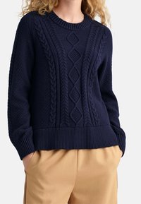 Granatowy dzianinowy sweter z teksturowanymi wzorami, warkoczowymi akcentami, prążkowanym dekoltem i mankietami, zestawiony z beżowymi spodniami.