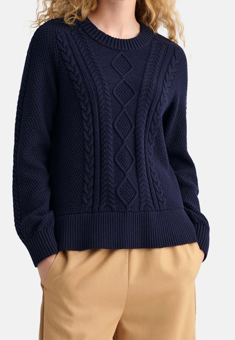 Granatowy dzianinowy sweter z teksturowanymi wzorami, warkoczowymi akcentami, prążkowanym dekoltem i mankietami, zestawiony z beżowymi spodniami.