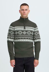 Pulover tricotat de culoare verde închis, cu guler înalt și fermoar până la jumătate, având modele geometrice albe și gri deschis pe piept și pe mâneci.
