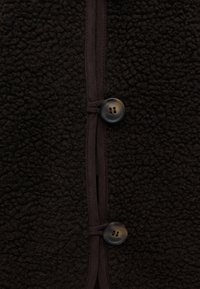 Gros plan sur un tissu polaire texturé marron foncé avec deux boutons marron foncé et une bordure assortie en ligne verticale.