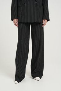 Completo a righe nere con giacca doppiopetto e pantaloni a gamba larga. Il tessuto ha una consistenza liscia, abbinato a scarpe bianche con punta.