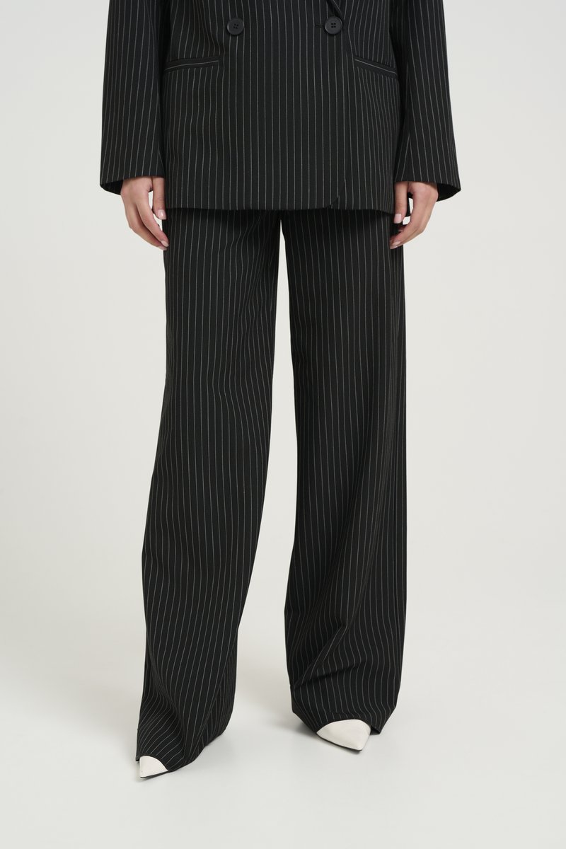 Completo a righe nere con giacca doppiopetto e pantaloni a gamba larga. Il tessuto ha una consistenza liscia, abbinato a scarpe bianche con punta.