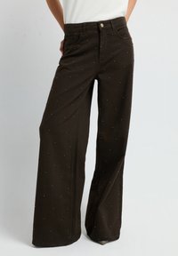 Pantalon large marron en coton, orné de clous métalliques décoratifs tout au long, avec une fermeture à boutons et des poches avant.