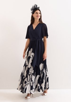 Femme portant une robe portefeuille bleu marine avec un grand imprimé floral blanc, des manches plissées, des talons blancs, une pochette assortie et un accessoire noir pour la tête.