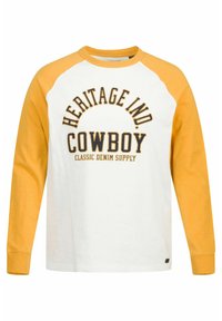 Langslavige t-shirt met een witte body, gele raglanmouwen en zwarte tekst: "HERITAGE IND. COWBOY CLASSIC DENIM SUPPLY."