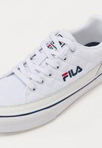 White canvas sneaker with quilted texture, round toe, and flat white laces. Features a blue and red FILA logo on the side and navy accents.