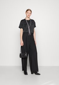 Schwarzes Satin-Top mit kurzen Ärmeln, kombiniert mit hochtaillierten, schwarzen, weiten Hosen. Eine silberne Halskette und eine schwarze, strukturierte Handtasche runden den Look ab.