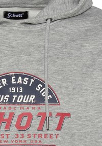 Sudadera gris con capucha, que presenta un parche de logo en azul marino y rojo. Incluye un cordón con puntas metálicas y una textura suave.