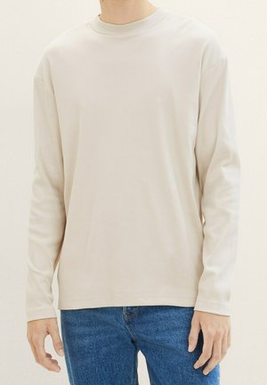 Personne portant une chemise à manches longues beige clair unie avec un jean bleu sur un fond neutre, vue du cou aux hanches.