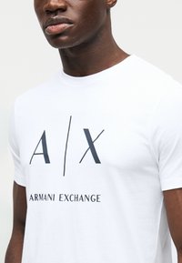 Tricou alb din bumbac cu un logo „A/X” albastru închis și textul „ARMANI EXCHANGE” centrat dedesubt. Mâneci scurte, gât rotund.