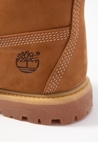 Botte en daim marron avec couture blanche, semelle en caoutchouc et logo d'arbre embossé. Présente une forme arrondie et une semelle extérieure texturée pour une meilleure adhérence.