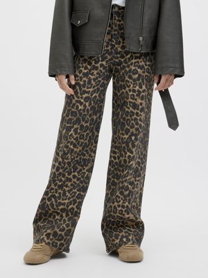 Persoană purtând pantaloni largi cu imprimeu leopard maro și negru, adidași tăbăciți și o geacă de piele neagră cu o curea care atârnă.
