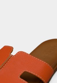 Oranžový kožený šľapák s textúrovaným povrchom, ktorý má výrezy a kontrastné biele prešívanie na vrchu. Hnedá vnútorná stielka.