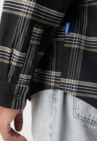 Schwarzes und olivfarbenes kariertes Flanellhemd mit weißen Akzenten, ausgestattet mit einem Knopfmanschette und über einem hellen Denimstoff getragen, der eine strukturierte Textur zeigt.