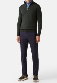 Hombre vestido con un suéter cerrado de color verde oscuro sobre una camisa azul, pantalones azul marino y zapatillas verde-marrón, de pie contra un fondo liso.