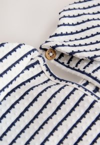 Primer plano de una tela blanca texturizada con rayas horizontales azul marino y un pequeño botón marrón que sujeta un cuello.