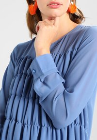 Blouse bleue avec texture froncée et poignets boutonnés. Tissu transparent, manches longues, col rond classique. Présente de grandes boucles d'oreilles en franges orange vif.