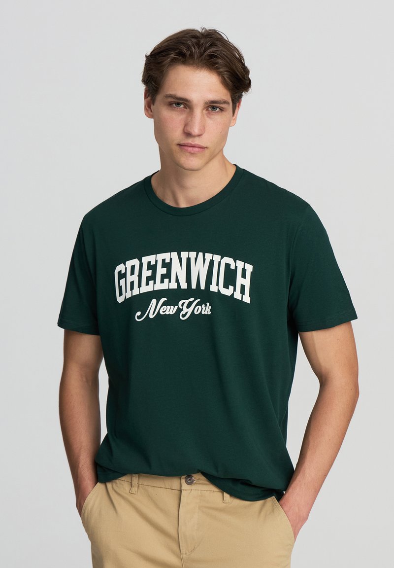 T-shirt verde scuro in cotone con scollatura rotonda. Presenta un testo bianco che recita "GREENWICH New York" in un design curvato.